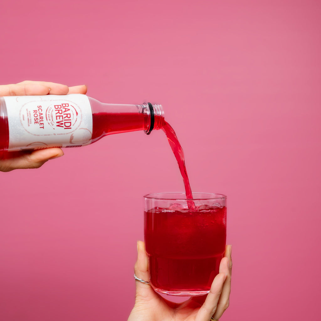 A hand pouring, a cold hibiscus tea in Kenya, that appears to be red from a bottle to a glass that's almost full. The bottle has a white label displaying the brand name and product name.